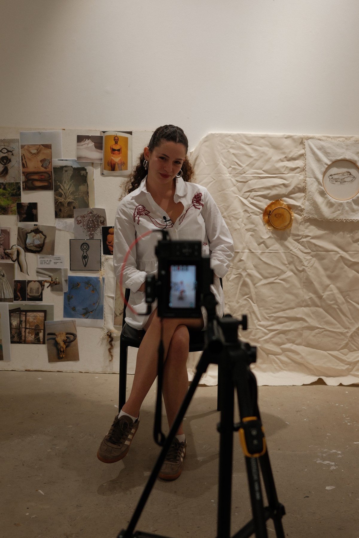 Jasmine Lane seated in her studio, framed by a wall of collaged research on one side and a hanging fabric installation with embroidery hoops on the other, camera and tripod in the foreground