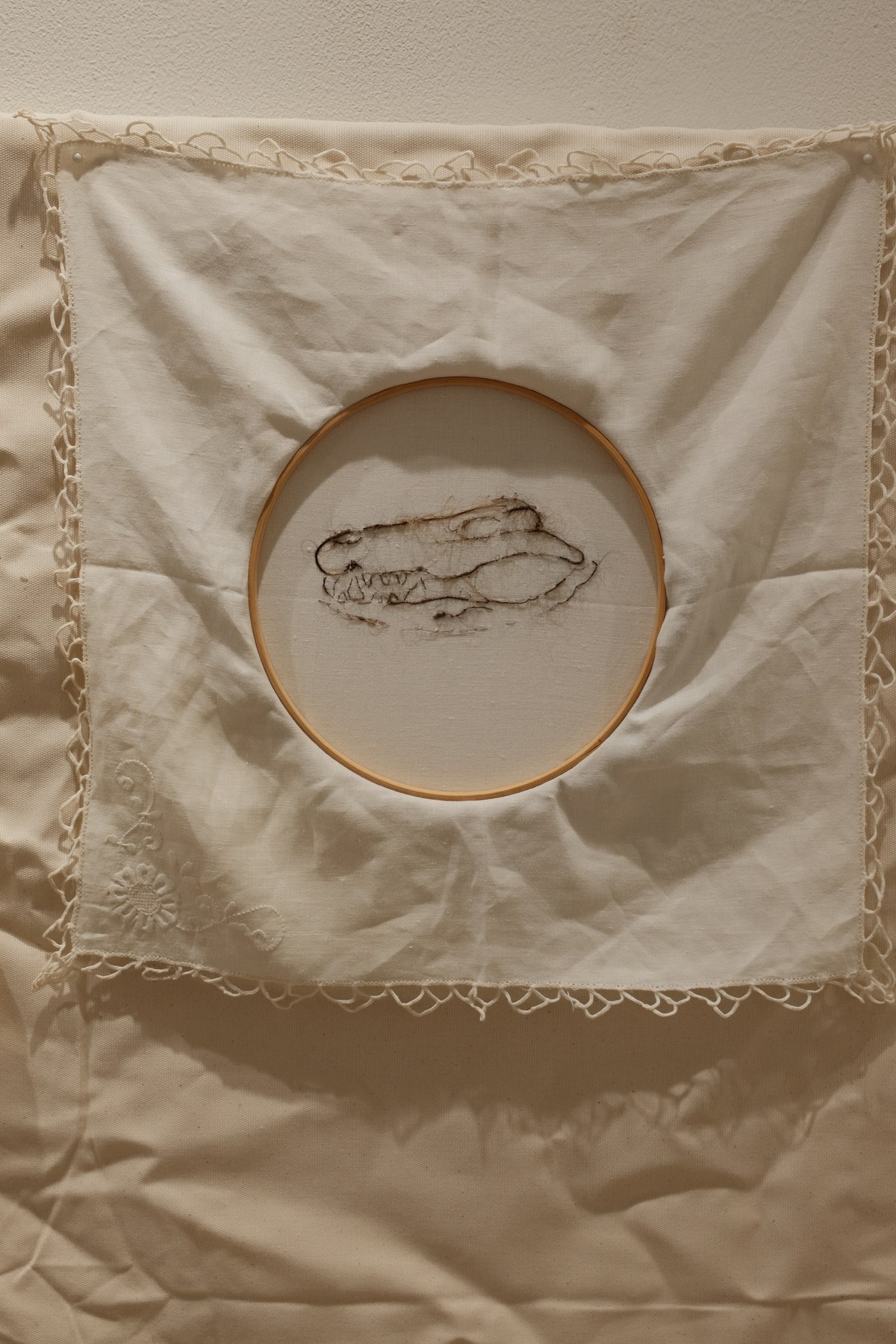 Close-up of an embroidered crocodile skull stitched in dark thread within a wooden hoop, framed by lace-edged white cloth