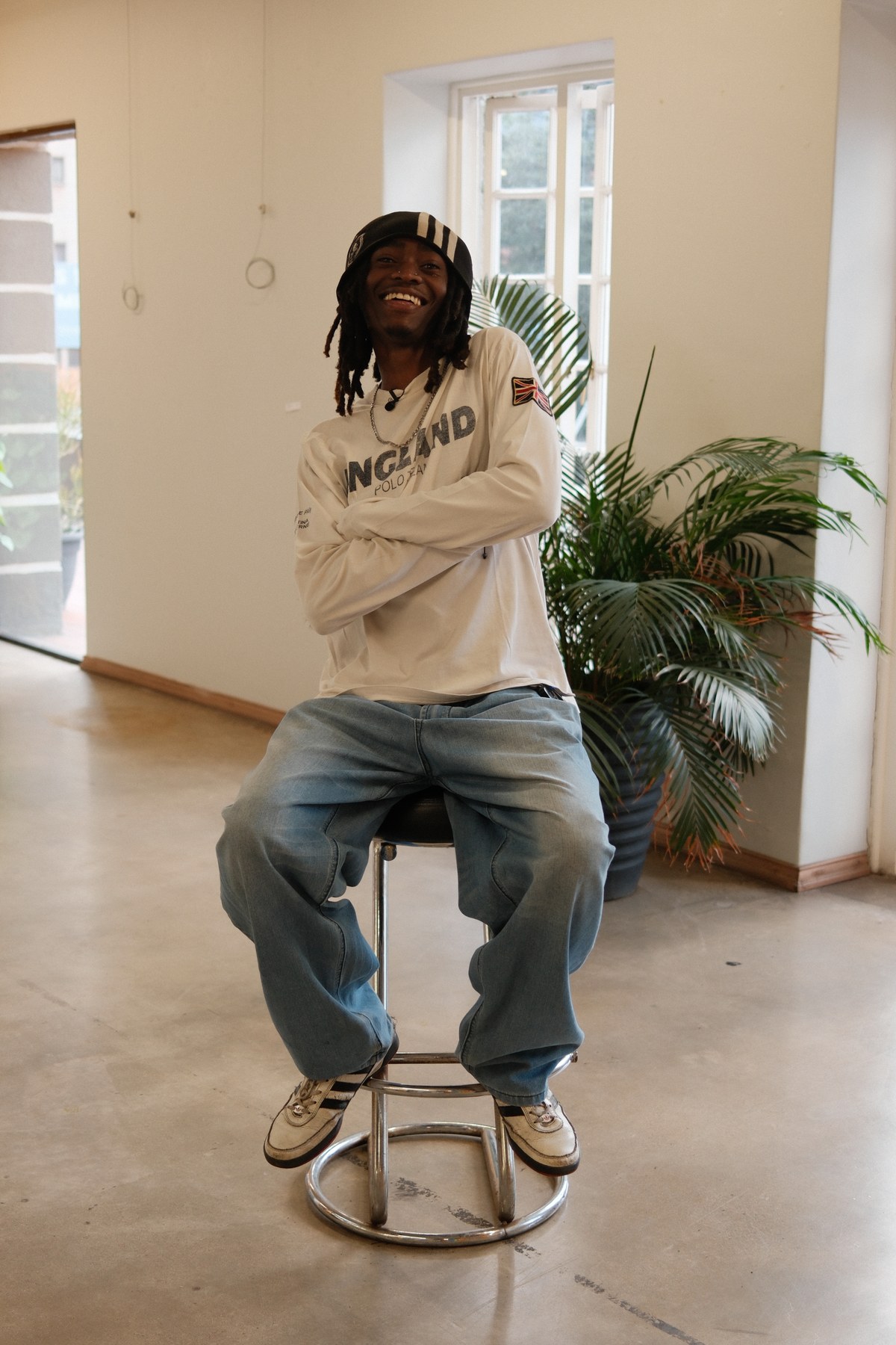 Kabelo Matsile seated on a stool in soft natural light, laughing, with a palm and window behind him