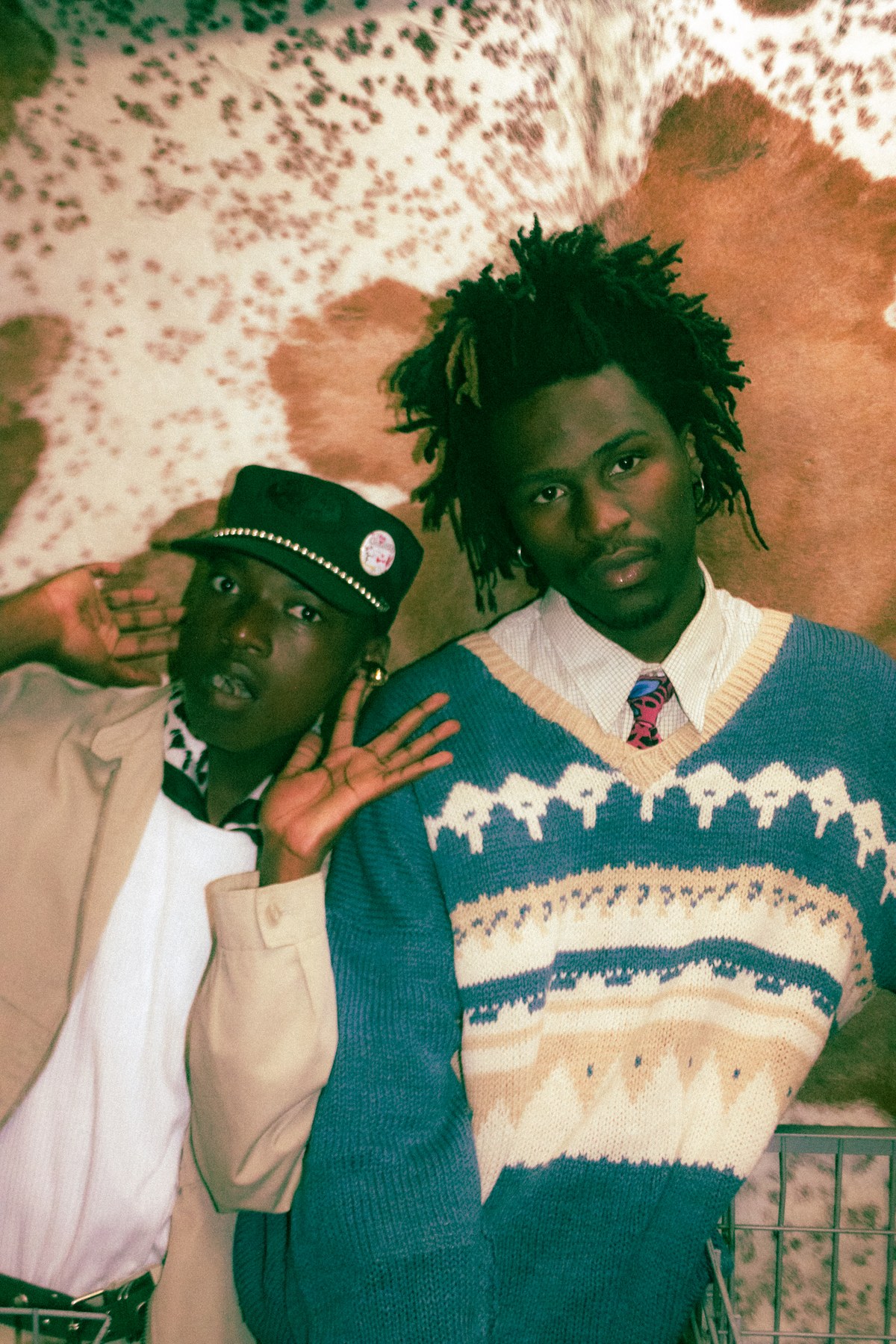 Photograph by Kabelo Matsile — double portrait against a cowhide print backdrop, one figure in a knit vest and tie, the other in a cap and white shirt