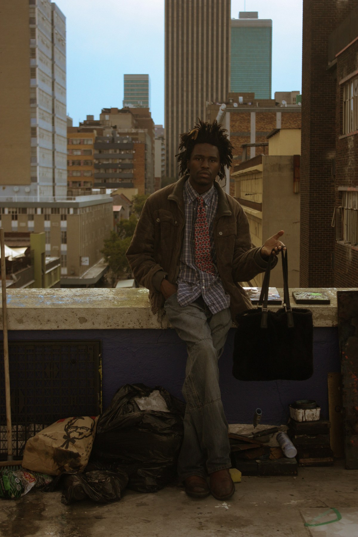 Photograph by Kabelo Matsile — figure leaning on a low wall on a Johannesburg rooftop, city skyline rising behind them
