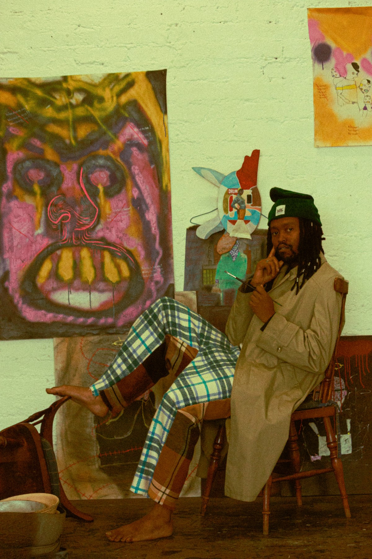 Photograph by Kabelo Matsile — portrait of an artist in a studio, seated beside a large expressive painted face on canvas