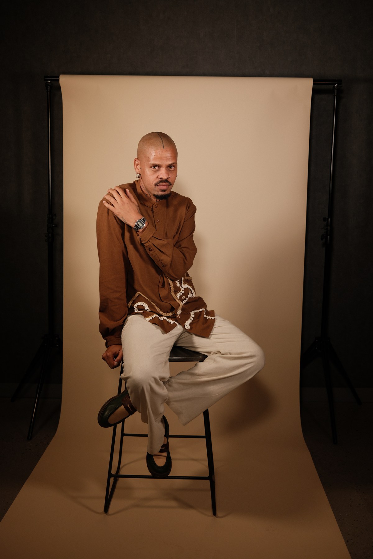 Kgosi Gift seated on a stool against a warm cream studio backdrop, wearing a brown shirt detailed with rope and cowrie embellishment, looking directly at camera