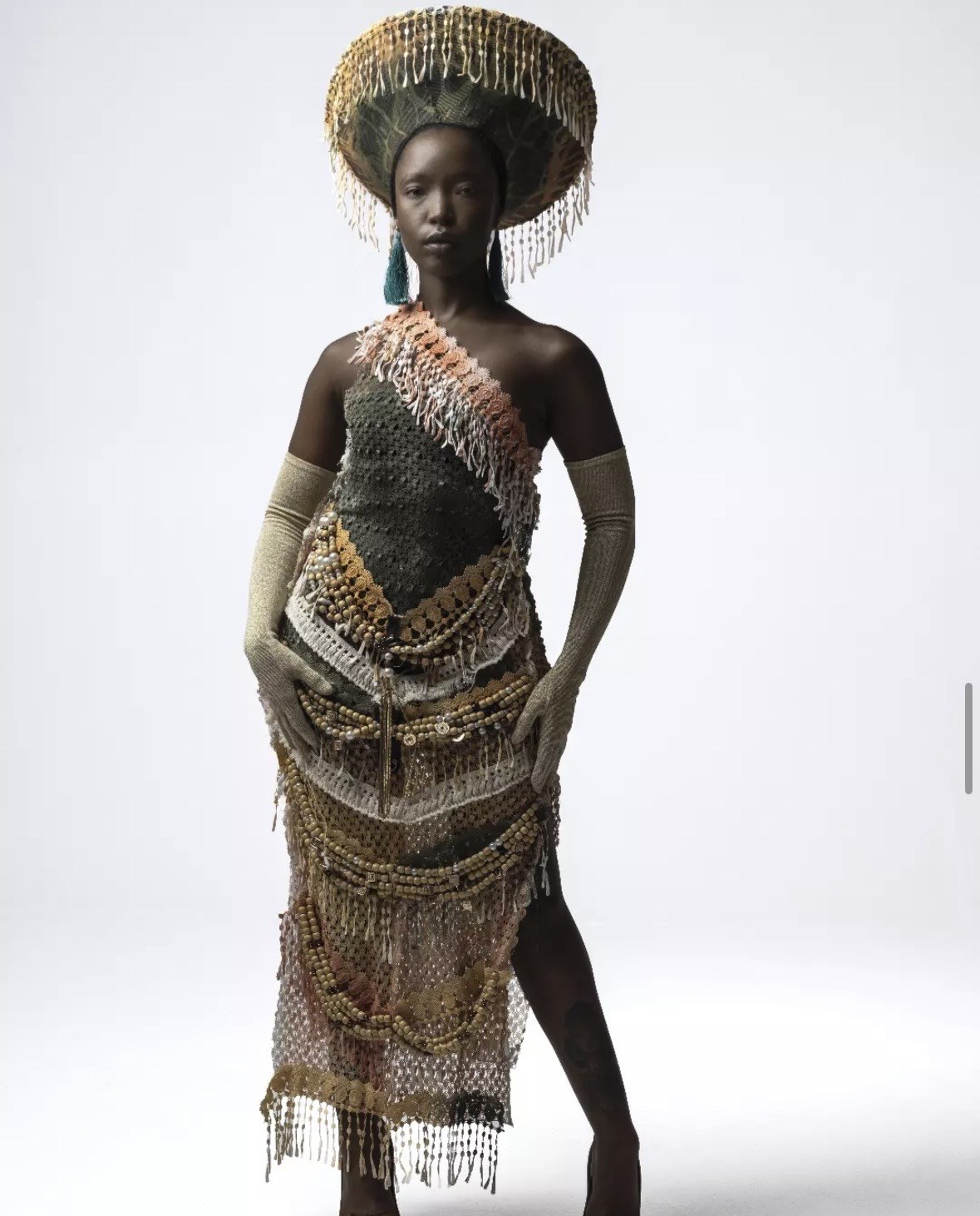 Designed by Kgosi Gift — a fringed beaded dress in olive, ivory, and ochre tones, worn with a wide tasselled headdress and long knit gloves