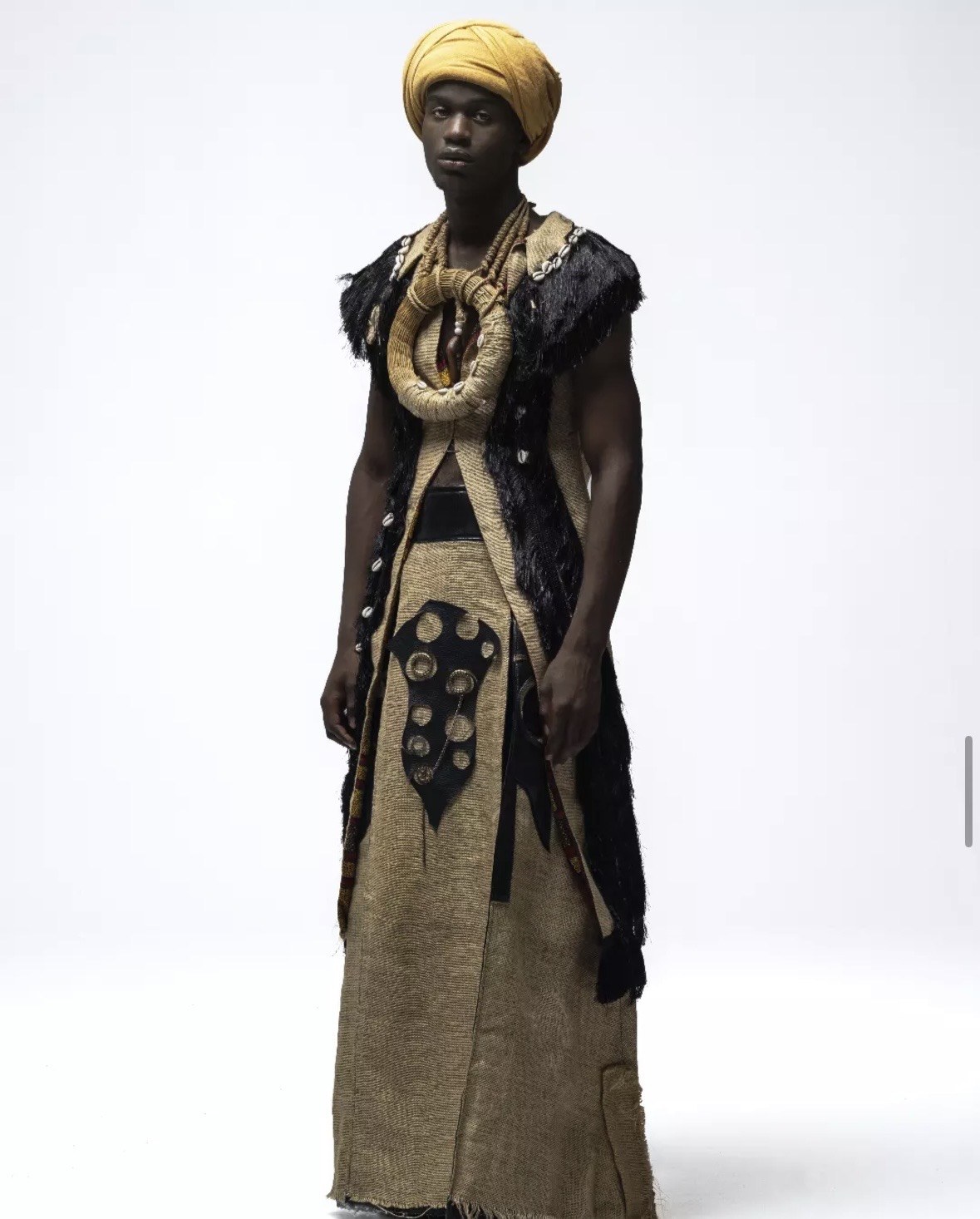 Designed by Kgosi Gift — a layered jute robe with a black fringed vest, woven cowrie neckpiece, and yellow headwrap