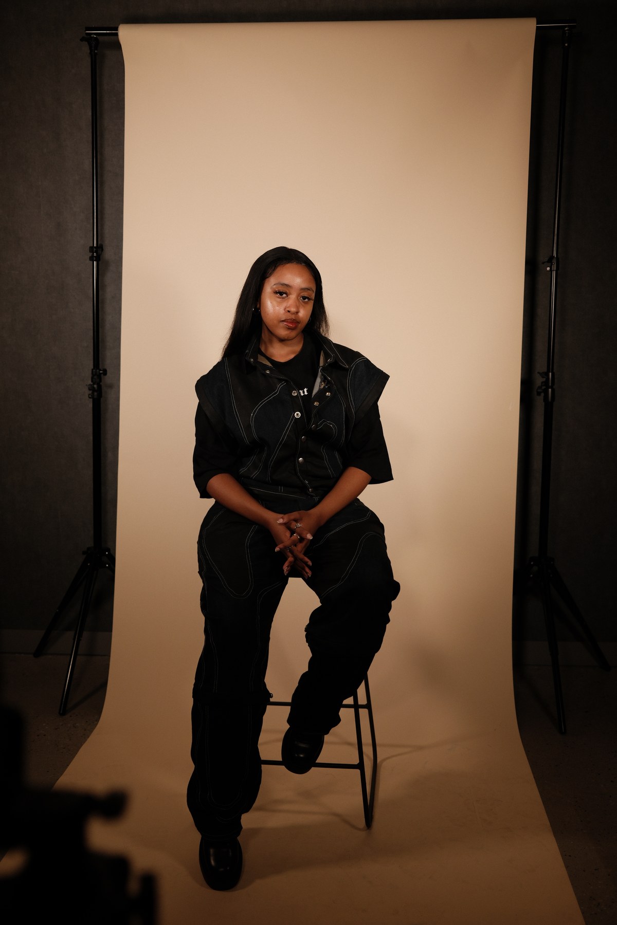 Mihlali Madikane seated on a stool against a warm cream studio backdrop, wearing a black denim look with contrast stitching from her own collection