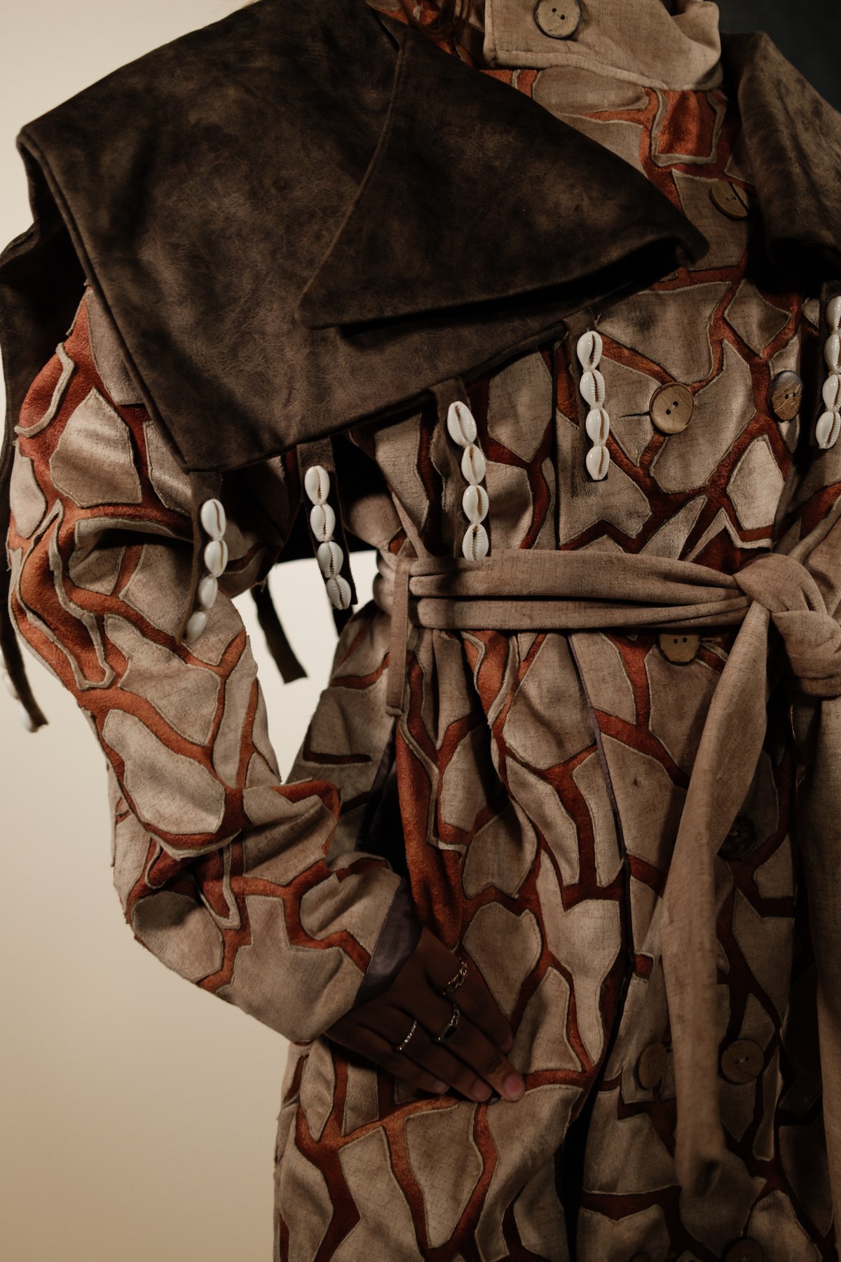 Designed by Mihlali Madikane — close detail of the patchwork jacket showing the leather hood, cowrie shell strands, and coconut buttons