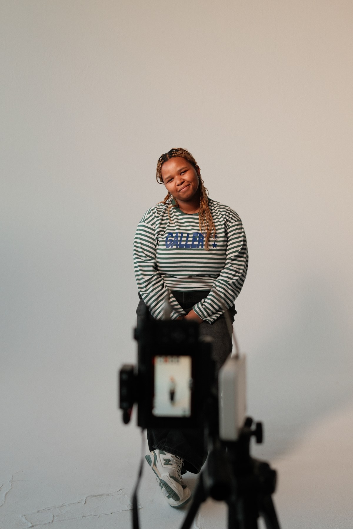 Oni Speelman in the studio, seated and smiling softly in a green striped long-sleeve, a camera and tripod in the foreground